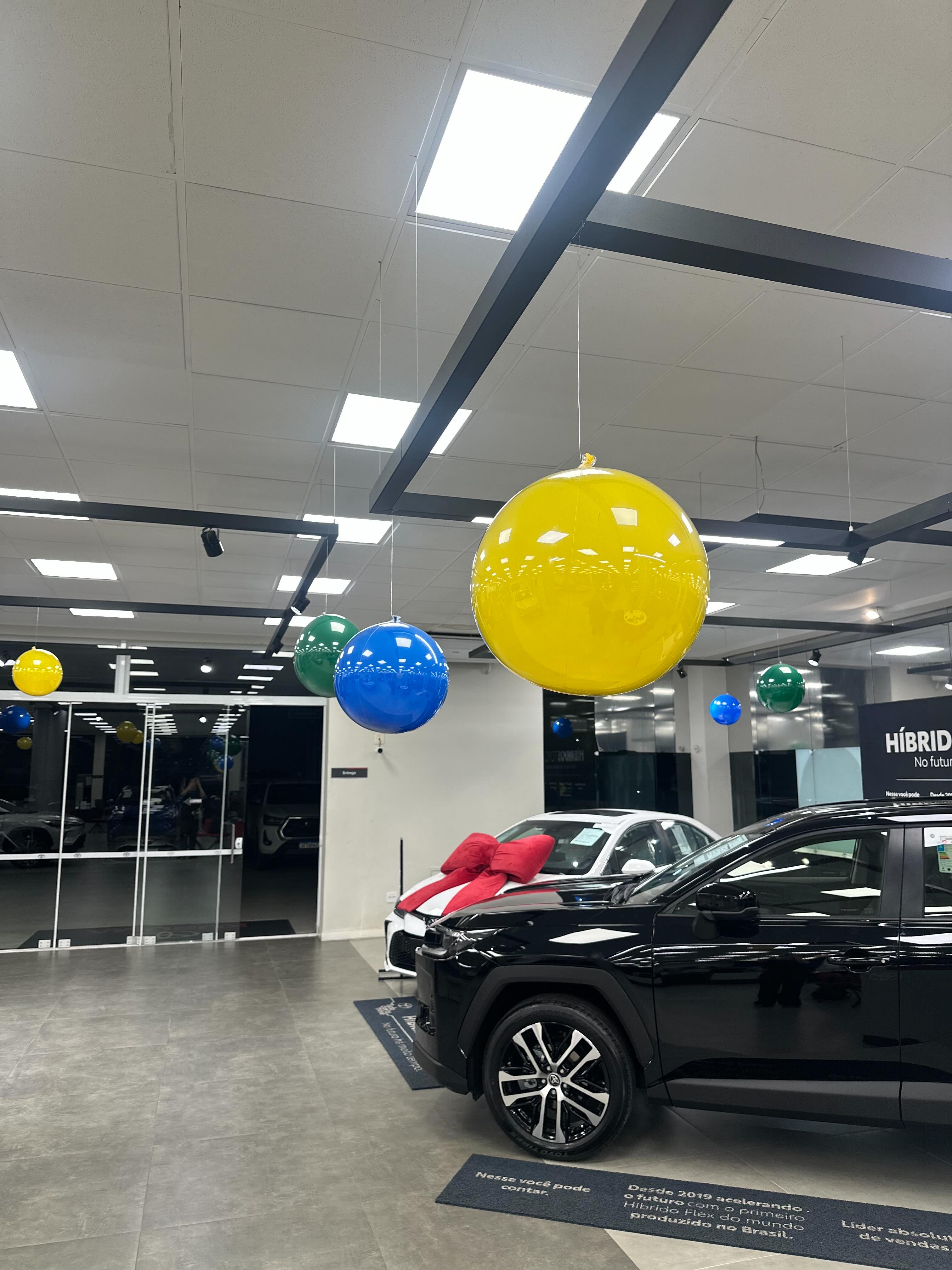 Balões amarelos, azuis e verdes suspensos sobre veículos em uma concessionária decorada para campanha especial.